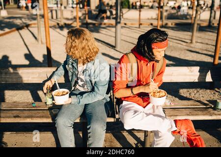 Gli amici che mangiano prendono il cibo mentre si siedono sulla panchina dentro città durante il giorno di sole Foto Stock