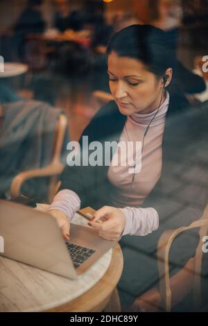 Vista ad alto angolo di una donna d'affari che fa acquisti online su un computer portatile mentre si siede in un caffè visto attraverso la finestra Foto Stock