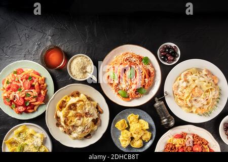 Cucina italiana. Molti diversi tipi di pasta, parmigiano grattugiato, ravioli, olive e vino, sparati dall'alto su fondo nero Foto Stock