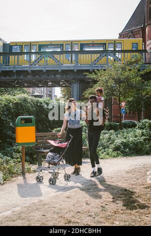 Padre che porta la figlia sulle spalle mentre la madre spinge il passeggino del bambino al sentiero contro il ponte ferroviario Foto Stock