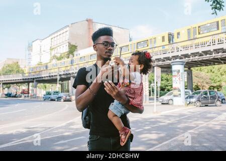 Giovane uomo che porta la figlia che tiene il telefono cellulare mentre si sta in piedi strada contro il ponte ferroviario Foto Stock