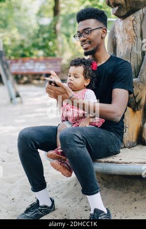 Bambina che usa il telefono cellulare mentre si siede con il padre sopra parco giochi all'aperto Foto Stock