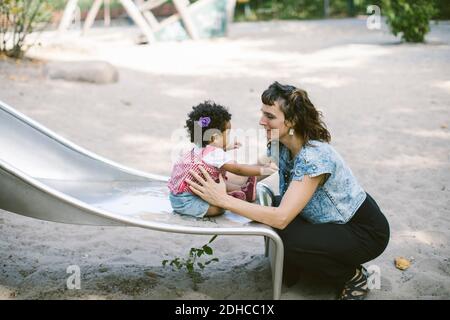 Vista laterale della madre che sostiene la figlia seduta sul vetrino a. parco giochi Foto Stock