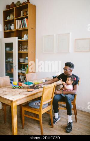 Giovane uomo che usa il computer portatile mentre si siede con la figlia sulla sedia al tavolo da pranzo in casa Foto Stock