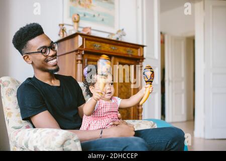 Bambina che tiene maracas mentre si siede con il padre felice sopra poltrona a casa Foto Stock