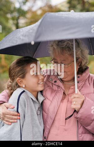 Nonna abbracciando la nipote mentre condivide l'ombrello con lei nel parco durante la stagione delle piogge Foto Stock