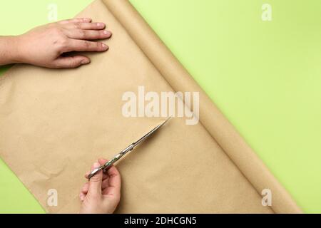 La mano femminile taglia un rotolo di carta marrone con il ferro forbici su sfondo verde Foto Stock