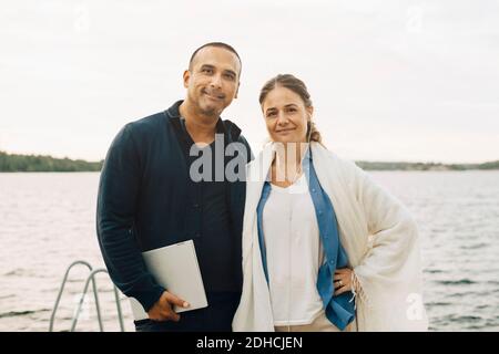 Ritratto di una coppia matura sorridente con un computer portatile in piedi contro il lago Foto Stock