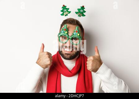 Natale, Capodanno e concetto di celebrazione. Primo piano di bell'uomo bearded in divertenti occhiali da festa che guarda felice, mostrando thum Foto Stock