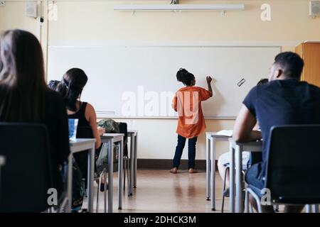 Vista posteriore della scrittura di un insegnante femminile sulla lavagna in classe Foto Stock