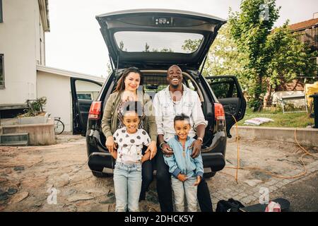 Ritratto di una famiglia multietnica sorridente appoggiata sul bagagliaio dell'auto cortile anteriore Foto Stock
