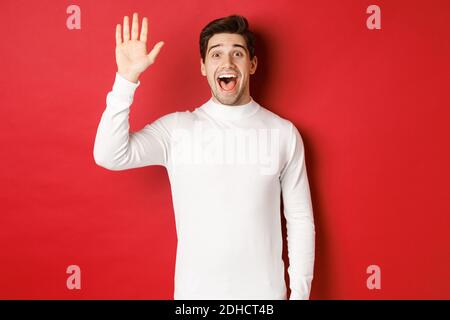 Immagine di un giovane felice e amichevole che saluta, agitando la mano per salutare qualcuno, in piedi su sfondo rosso Foto Stock