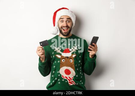 Concetto di shopping online e vacanze invernali. Uomo sorpreso in cappello di Santa, tenendo il telefono cellulare e la carta di credito, in piedi nel sudore Foto Stock