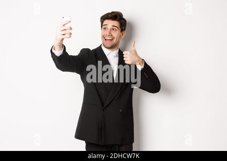 Ritratto di un uomo dall'aspetto bello che prende selfie durante la festa del nuovo anno, indossa il vestito, scatta foto sullo smartphone e mostra i pollici, st Foto Stock