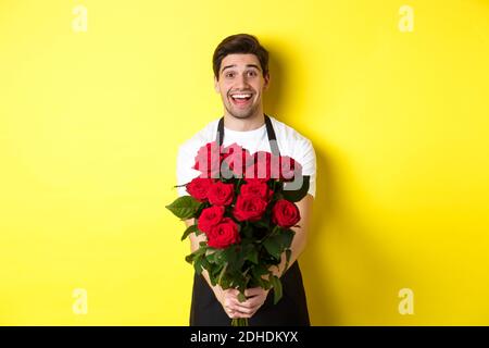 Venditore in negozio di fiori che indossa grembiule nero, dando bouquet di rose e sorridente, in piedi su sfondo giallo Foto Stock