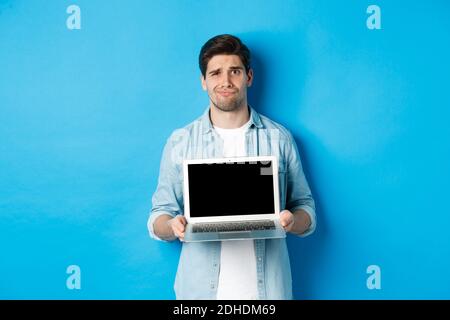 Un ragazzo oscuro e dispiaciuto che mostra lo schermo del notebook e la grimacing, avendo dubbi, in piedi su sfondo blu in casual Foto Stock