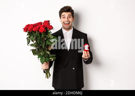 Bell'uomo sorridente in tuta nera, con le rose e l'anello di fidanzamento, facendo una proposta di sposarlo, in piedi contro la ba bianca Foto Stock