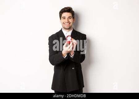 Immagine di bel ragazzo in vestito nero facendo una proposta, chiedendo di sposarlo e mostrando anello di nozze, in piedi su bianco b Foto Stock