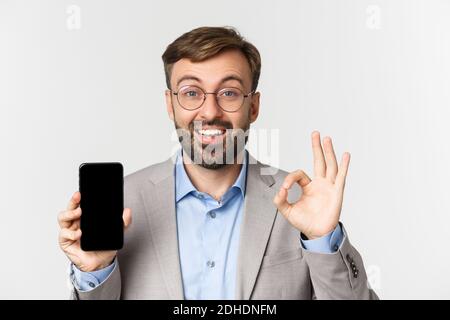 Primo piano di un uomo d'affari entusiasta in abito grigio e occhiali, raccomandando una buona app, mostrando lo schermo del telefono cellulare e OK segno, stan Foto Stock
