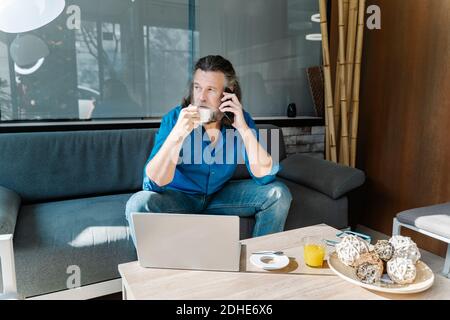 Uomo maturo con la barba che parla al telefono e beve un caffè seduto su un divano di fronte al portatile che guarda fuori dalla finestra. concetto di business Foto Stock