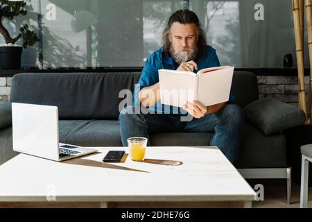Uomo maturo con una barba che legge un libro seduto sopra un divano nel soggiorno Foto Stock