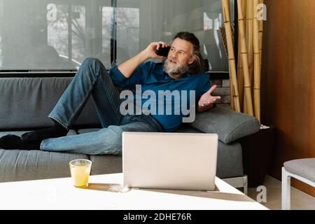 Uomo maturo con la barba che parla al telefono su un divano di fronte al computer portatile. concetto di business Foto Stock