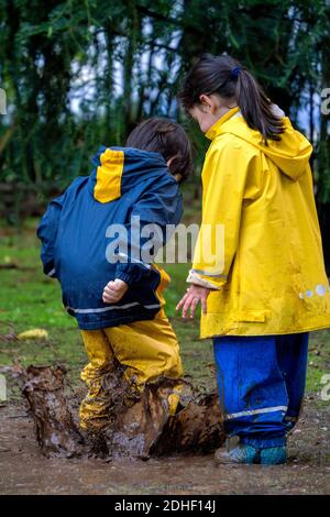 Due bambini piccoli che si divertono a giocare fuori saltando in un fango puddle indossando gli iridati coloful Foto Stock