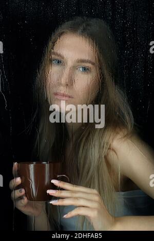 Blocco. Ritratto di giovane bionda con tazza di caffè in mano. Femmina dietro il vetro del finestrino con gocce di pioggia. Foto Stock