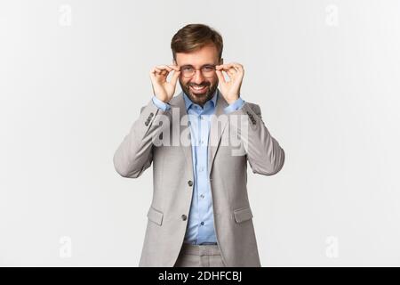 Ritratto di riuscito uomo d'affari bearded in vestito grigio, messo sugli occhiali e sorridente soddisfatto, in piedi su sfondo bianco Foto Stock