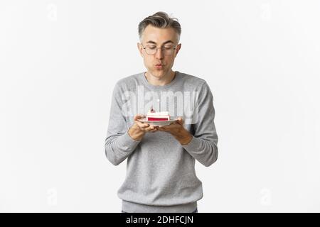 Ritratto di attraente uomo di mezza età in pullover grigio e bicchieri, celebrando il suo compleanno, soffiando una candela su torta b-day e. Foto Stock