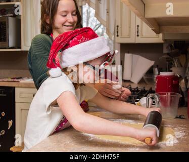 Giovane ragazza che usa il perno di rotolamento per fare i biscotti dello zucchero per Natale come la sorella grande aiuta. Indossare un cappello e occhiali da vacanza, mentre si utilizza il perno di rotolamento Foto Stock