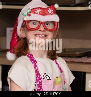 Giovane ragazza sorridente in cucina con cappello santa e occhiali da vacanza con grembiule rosa. Foto Stock