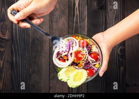 Vista dall'alto delle mani della donna che tengono in mano la ciotola per insalata colorata e sana con verdure miste fresche in un piatto su sfondo di legno nero, snack Salute f Foto Stock