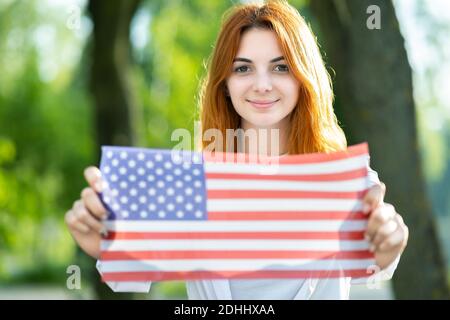 Felice giovane donna in posa con la bandiera nazionale degli Stati Uniti che la tiene nelle sue mani distese in piedi all'aperto nel parco estivo. Ragazza positiva che celebra Unite Foto Stock