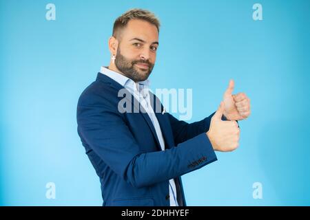 Giovane uomo che indossa il costume con i pollici su sfondo blu. Foto Stock