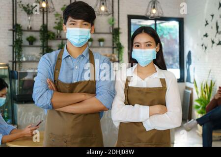 Coppia barista asiatica in grembiule e maschera facciale guardando Macchina fotografica pronta per offrire il servizio caffè al caffè moderno negozio Foto Stock