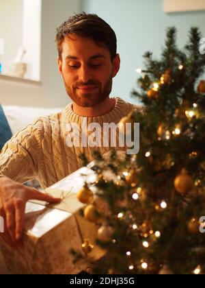 Giovane caucasico accanto ad un albero di Natale in un maglione di lana che tiene un regalo nelle sue mani. Gode le vacanze in una casa accogliente. Foto Stock