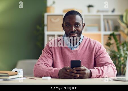 Ritratto di un giovane africano sorridente alla macchina fotografica mentre è seduto al tavolo e utilizzando il suo cellulare Foto Stock