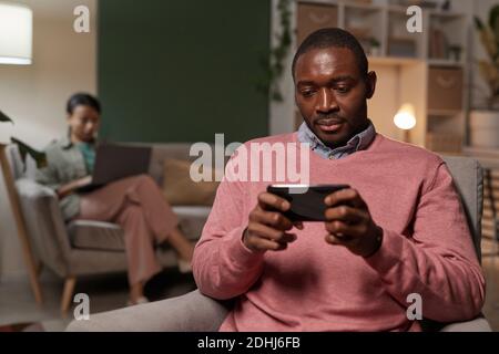 Giovane africano seduto sulla poltrona e giocando il suo telefono cellulare con la donna che usa il computer portatile in background Foto Stock