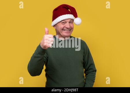 Uomo anziano che indossa il cappello di babbo natale facendo i pollici felici su gesto con la mano. Approvazione dell'espressione guardando la videocamera. Foto Stock