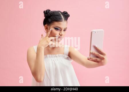 Cute sorridente ragazza asiatica adolescente prendendo auto shot foto con la sua fotocamera del telefono, su sfondo rosa Foto Stock