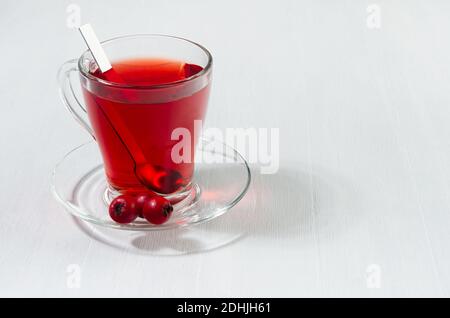 Tè rosso in elegante tazza trasparente con bacche di biancospino mature alla luce del sole su tavola di legno bianco, spazio copia. Foto Stock