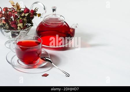 Colazione con tè biologico rosso biancospino in teiera trasparente, tazza, frutti di bosco biancospino, rosa anca alla luce del sole con ombra su tavola di legno bianco, copia spac Foto Stock