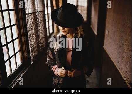 Elegante donna in elegante abito vintage e cappello in piedi stretto corridoio della vecchia casa e guardando lo specchio Foto Stock