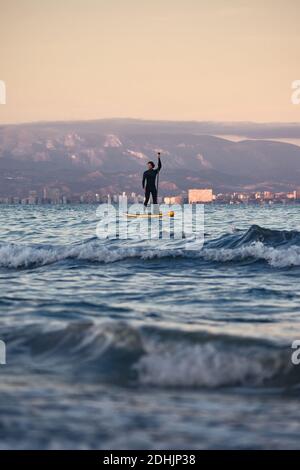 Surfista maschile in muta canottiera su tavola a pale in mare acqua sullo sfondo delle montagne al tramonto Foto Stock