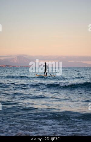 Surfista maschile in muta canottiera su tavola a pale in mare acqua sullo sfondo delle montagne al tramonto Foto Stock