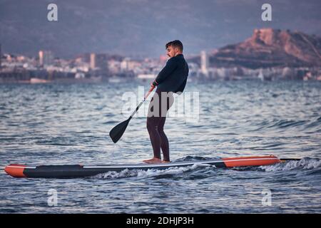 Vista laterale del surfista maschile in muta canottaggio sulla pagaia sali sull'acqua di mare sullo sfondo delle montagne al tramonto Foto Stock
