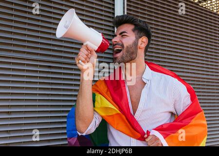 Da sotto eccitato omosessuale maschio in piedi vicino a costruzione con LGBT bandiera arcobaleno e gridando in altoparlante Foto Stock