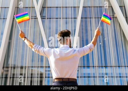 Vista posteriore basso angolo di uomo omosessuale in piedi sulla strada Con bandierine LGBT di carta e guardando via Foto Stock