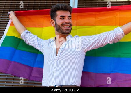 Positivo gay maschio in piedi con arcobaleno bandiera LGBT in città e guardando via Foto Stock